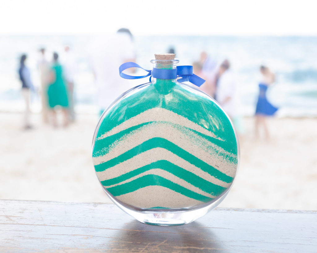 Turquoise and tan sand jar created by the bride & groom with guests in the background.