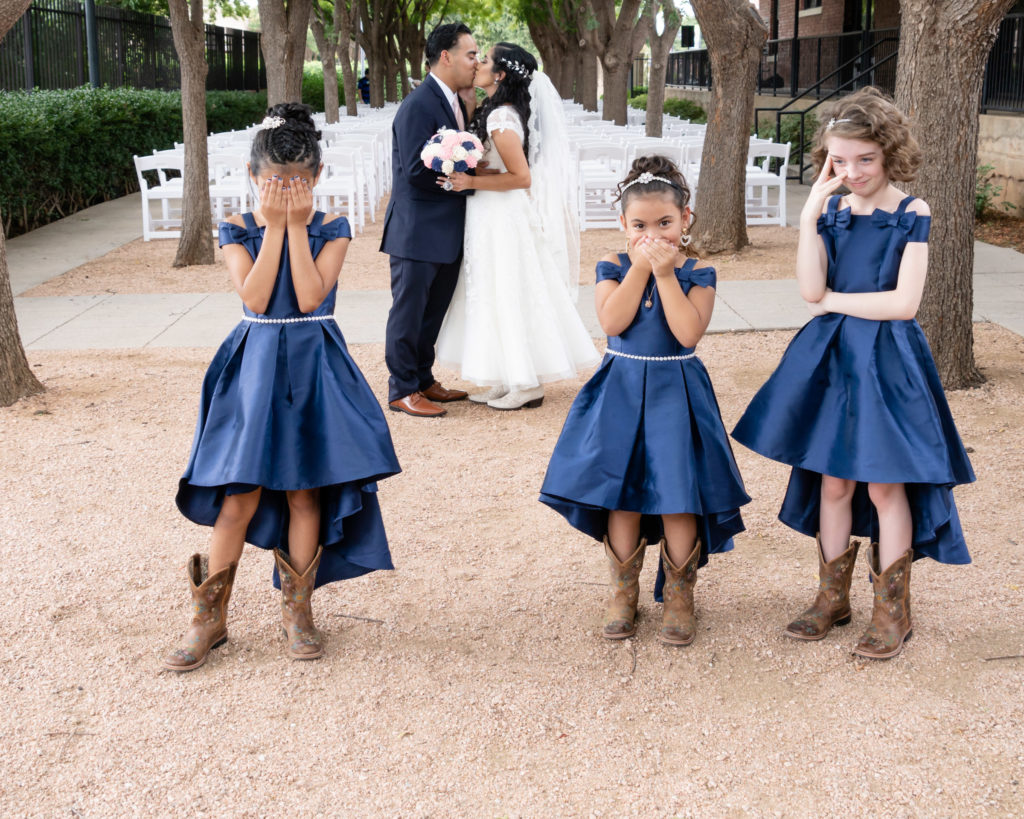 Latino bride & groom kissing in the background. Three flower girls in blue dresses in front. One has hands over her eyes, the middle with hands over her mouth, and one on right is smiling in embarrassment.