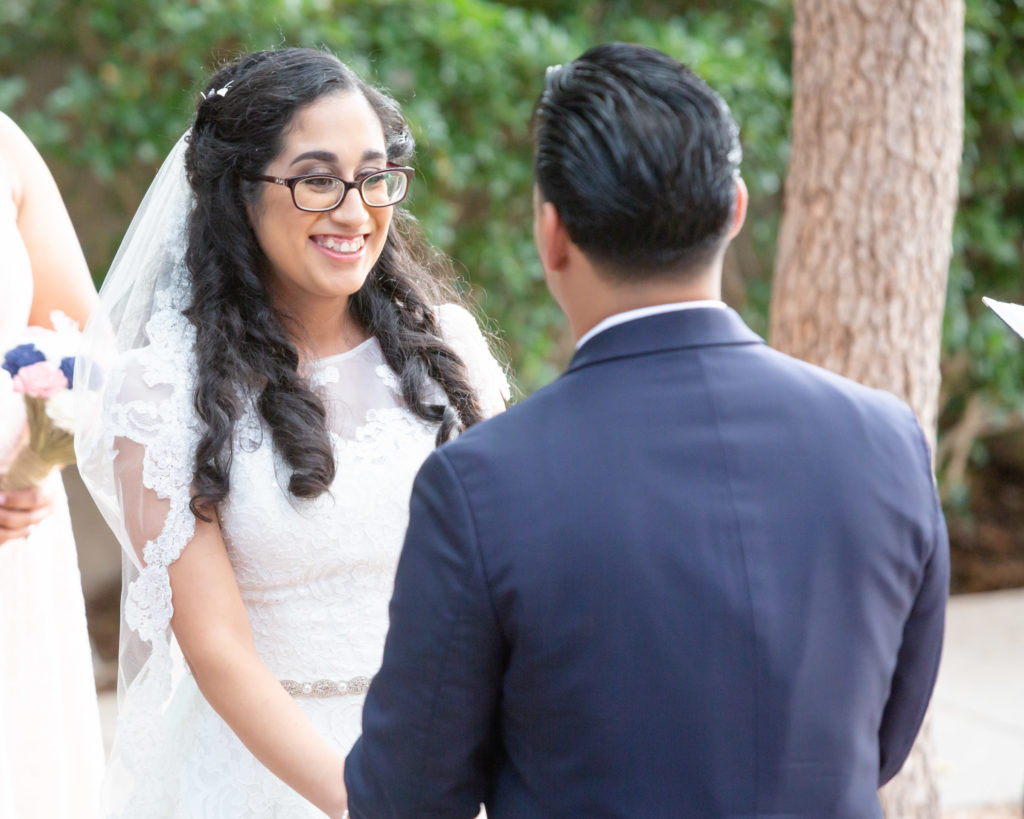 Front of Latina bride smiling and facing the groom. Back of groom in a blue jacket.