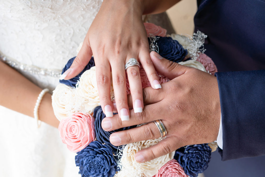 Bouquet with white, pink, and blue flowers. Bride & Grooms hands with rings are on top of bouquet.