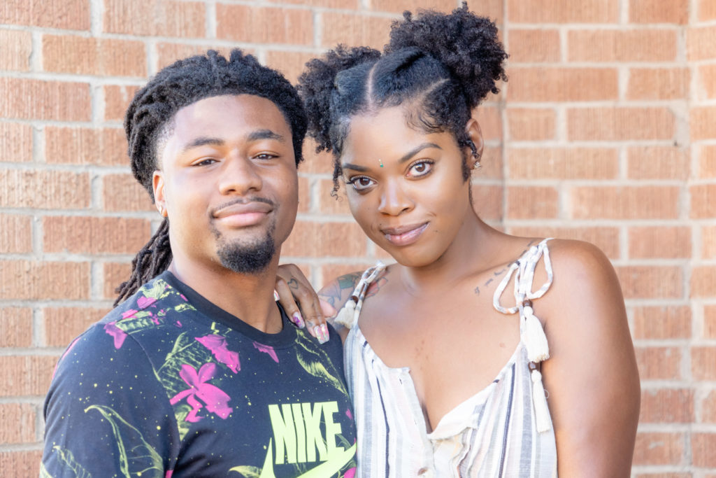 African American couple. Headshot of them with her arm behind him resting her hand on his shoulder. He's in a black Nike shirt. She's in a white strappy dress with her hair in pigtails. Dallas photographer