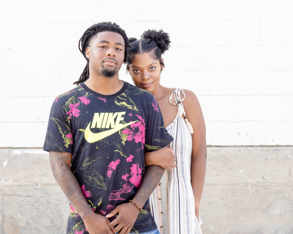 African American couple. He's in a black Nike shirt with pink flowers. She's in a striped gray, white, and pink dress with her hair braided into pigtails. Slightly smiling. She's half behind him wither her arm tucked around his elbow. Dallas photographer
