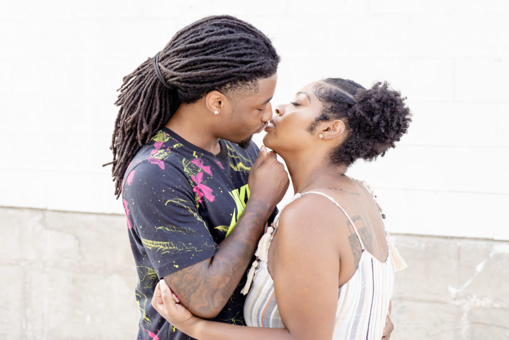 African American couple. He's in a black Nike shirt with pink flowers. She's in a striped gray, white, and pink dress with her hair braided into pigtails. Slightly smiling. Her arms are around him and his hand is tilting her chin up so they can kiss. Dallas photographer