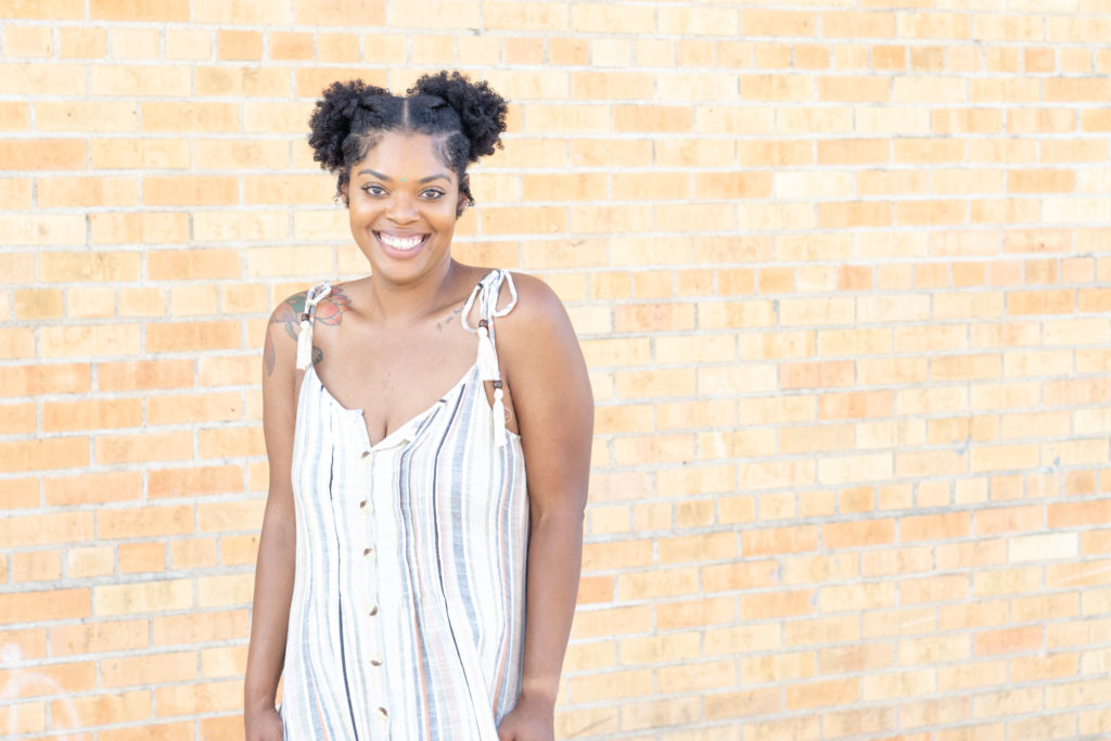 Beautiful African American woman in a striped gray, black, and white dress with  tied straps and her hair braided into pigtails. 3/4 length shot. She's smiling at the camera. Dallas photographer